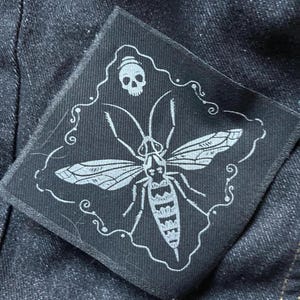 May include: Black and white embroidered patch featuring a moth with a skull above it. The moth has a detailed design and is surrounded by a decorative border.