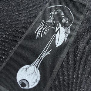 May include: A black and white illustration of a crow holding an eyeball in its beak. The crow is depicted in a detailed, stylised manner, with its feathers and beak clearly visible. The eyeball is large and prominent, with a visible iris and pupil.