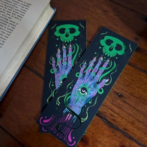 May include: Two black bookmarks with a spooky design. Each bookmark features a green skull at the top and a purple hand with an eye in the palm. The hand has long, pointed nails and is surrounded by green, swirling lines. The design is printed on a dark background.