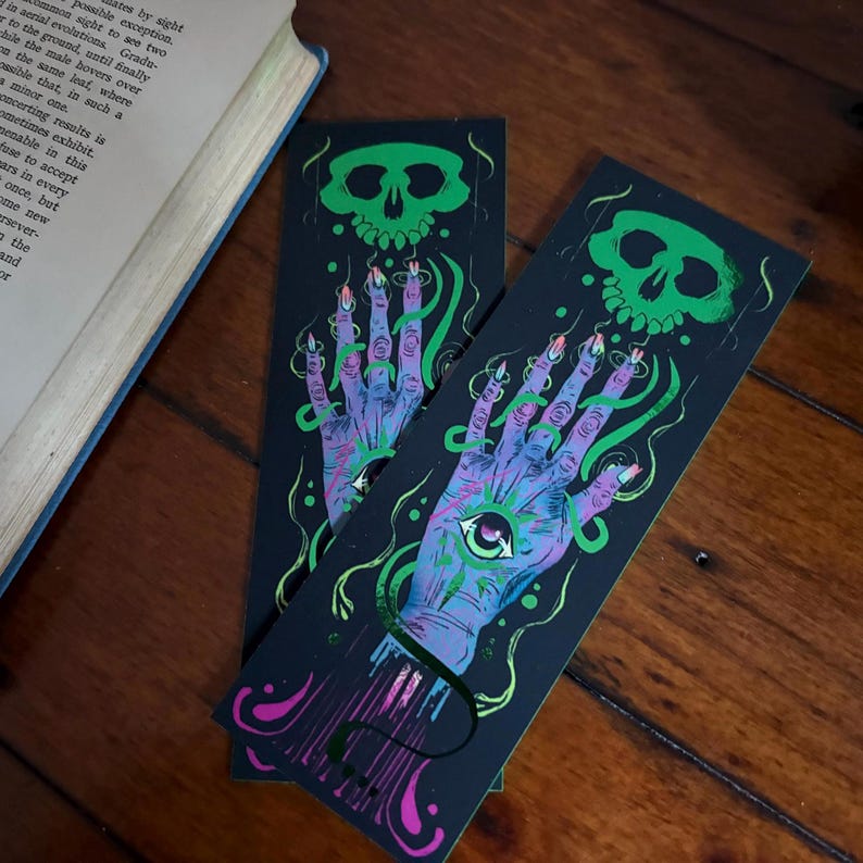 May include: Two black bookmarks with a spooky design. Each bookmark features a green skull at the top and a purple hand with an eye in the palm. The hand has long, pointed nails and is surrounded by green, swirling lines. The design is printed on a dark background.