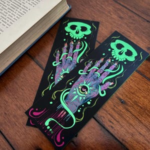 May include: Two black bookmarks with a spooky design featuring a glowing green skull, a hand with an eye in the palm, and swirling green and pink accents. The bookmarks are on a wooden surface.