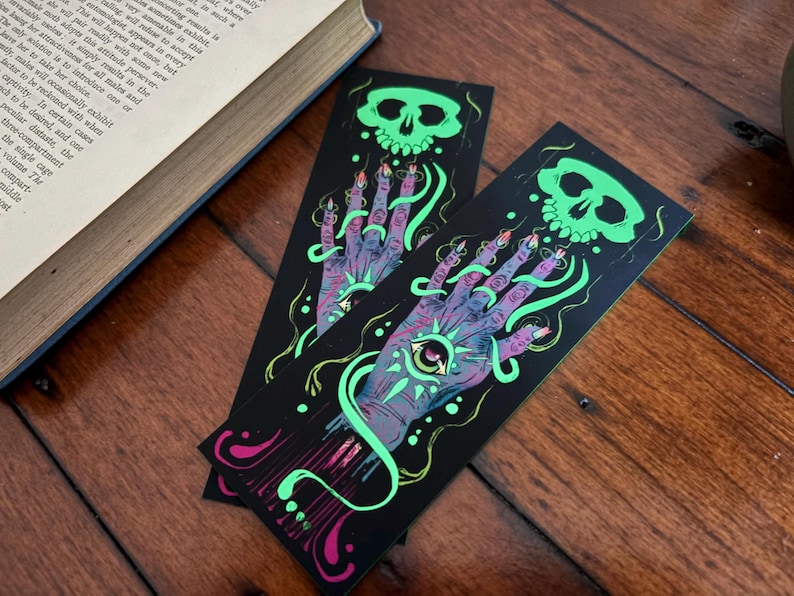 May include: Two black bookmarks with a spooky design featuring a glowing green skull, a hand with an eye in the palm, and swirling green and pink accents. The bookmarks are on a wooden surface.
