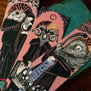 Può includere: Tre segnalibri rosa con illustrazioni in bianco e nero di creature fantastiche. Ogni segnalibro presenta una creatura diversa, tra cui un ariete, un uccello e una creatura con occhi grandi. I segnalibri hanno dettagli in oro e sono decorati con motivi floreali.