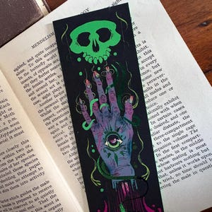 May include: Black bookmark with a green skull and a hand with an eye in the palm. The hand is purple and green with long fingernails. Green vines and drips of pink and purple are also present. The bookmark is next to an open book.