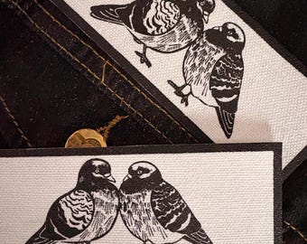 May include: Two rectangular fabric patches featuring a black and white screen-printed illustration of two pigeons standing side by side. The patches have a textured white surface with a solid black border, showcasing detailed linework.