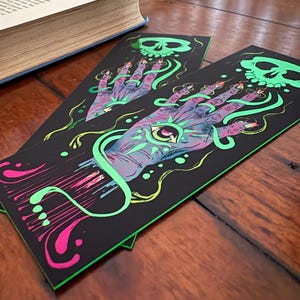 May include: Two black bookmarks with a neon green and pink design. The design features a hand with an eye in the palm, a skull, and swirling lines. The bookmarks are on a wooden surface next to a book.