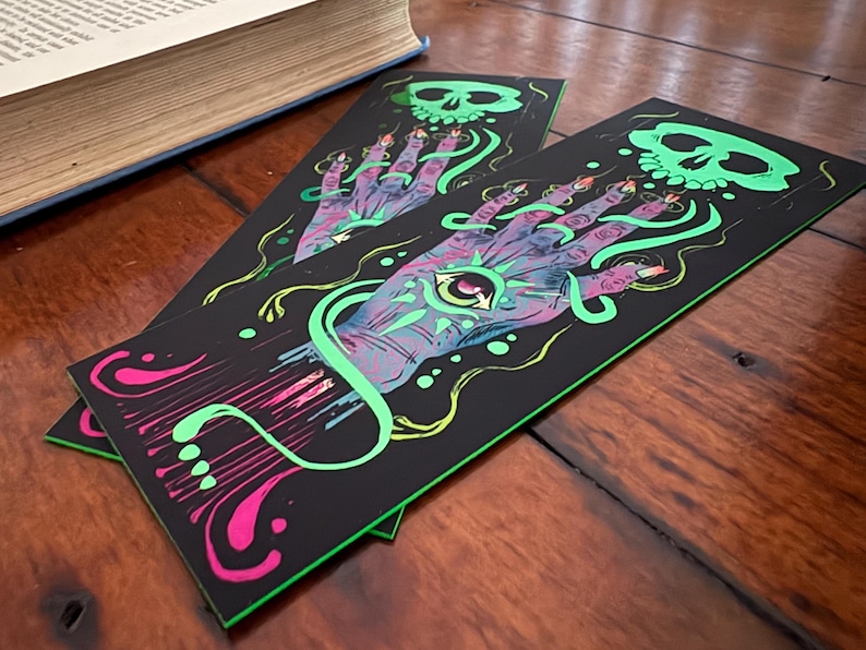 May include: Two black bookmarks with a neon green and pink design. The design features a hand with an eye in the palm, a skull, and swirling lines. The bookmarks are on a wooden surface next to a book.
