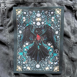 May include: A black patch with a detailed illustration of a raven with outstretched wings, holding a red heart. The design includes teal and white floral accents and an eye symbol, set against a dark background.