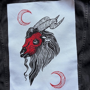 Può includere: Una toppa bianca con un'illustrazione nera e rossa di una testa di capra e mezzelune. La capra ha lunghe corna ricurve e una maschera rossa. La toppa è cucita su una giacca di jeans nera.