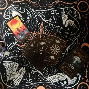 May include: A black tarot cloth with a white and gold design featuring birds, stars, and moons. A deck of tarot cards is spread out on the cloth, including the "The Sun" card. A small white bag with a blue stone is in the lower left corner. The text "Occult Ornithology" is visible on a black book in the lower right corner.