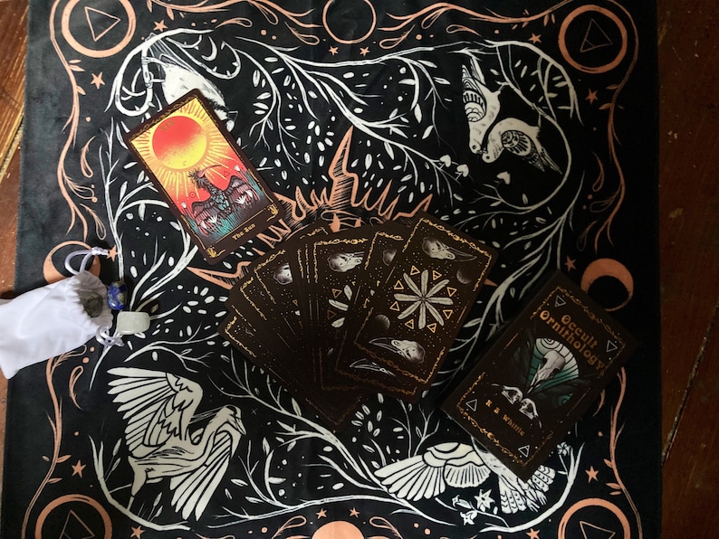 May include: A black tarot cloth with a white and gold design featuring birds, stars, and moons. A deck of tarot cards is spread out on the cloth, including the "The Sun" card. A small white bag with a blue stone is in the lower left corner. The text "Occult Ornithology" is visible on a black book in the lower right corner.