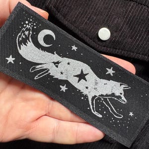 May include: A rectangular black patch featuring a silver fox leaping across a starry night sky. The fox is detailed with stars, a crescent moon, and other celestial elements. The patch is held against a black corduroy fabric.