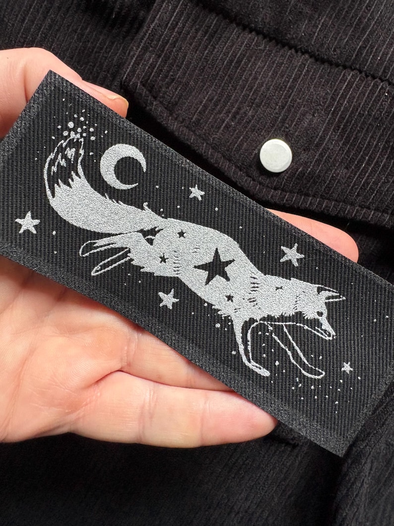 May include: A rectangular black patch featuring a silver fox leaping across a starry night sky. The fox is detailed with stars, a crescent moon, and other celestial elements. The patch is held against a black corduroy fabric.