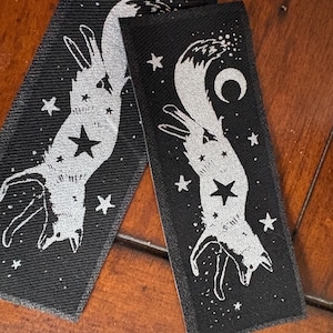 May include: Two black rectangular patches with a silver fox design, a star, and other celestial elements. The fox is upside down, surrounded by stars and a crescent moon. The patches are on a wooden surface.