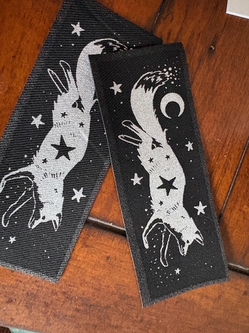 May include: Two black rectangular patches with a silver fox design, a star, and other celestial elements. The fox is upside down, surrounded by stars and a crescent moon. The patches are on a wooden surface.