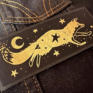Könnte beinhalten: Ein rechteckiger, schwarzer Aufnäher mit einem goldenen Fuchs, der über einen Nachthimmel mit Sternen springt. Das Design umfasst einen Mond und Sterne. Geeignet für Kleidung oder Accessoires.