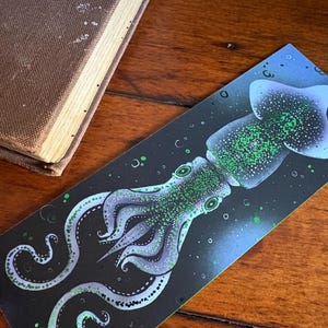 May include: A rectangular bookmark featuring a stylized illustration of a squid against a dark blue and black background. The squid is depicted in shades of green and purple, with detailed tentacles and eyes. Bubbles are scattered around the squid.