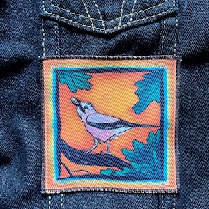 May include: A colourful embroidered patch featuring a pink and white bird perched on a branch with green leaves against an orange background.