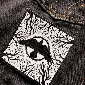 Könnte beinhalten: Quadratischer schwarz-weißer Aufnäher mit einem fliegenden Raben in einem Kreis, umgeben von einem Astdesign. Der Aufnäher befindet sich auf einem dunklen Jeanshintergrund mit sichtbaren gelben Nähten.
