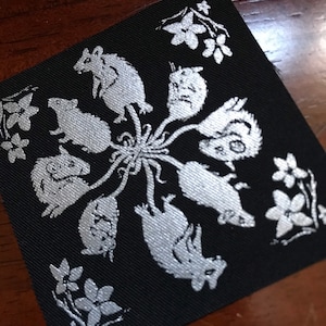 May include: A black fabric patch with a silver design of eight mice in a circle around a central flower. The mice are facing outward and have long tails that intertwine in the center.