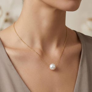 May include: A single white spherical pearl pendant suspended on a delicate gold chain necklace. The smooth, lustrous pearl is centered on the thin metallic chain, which rests against the collarbone area, showcasing a simple and classic design.