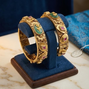 Puede incluir: Dos brazaletes articulados de tono dorado con un intrincado diseño floral texturizado. Cada brazalete está adornado con piedras cabujón ovaladas de color verde con motivos florales en relieve y piedras rojas cuadradas en marcos metálicos.