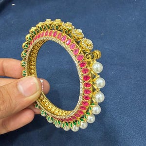 Pode incluir: Uma pulseira dourada com uma fileira de pedras cor-de-rosa em forma de gota, detalhes em esmalte verde e uma borda de pequenas pérolas brancas. A superfície metálica apresenta padrões intrincados e pequenas pedras claras na borda interna.
