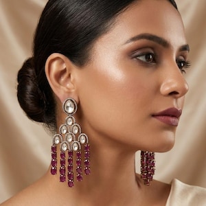 May include: A pair of chandelier earrings featuring a dark metal setting with clear, oval-shaped stones at the top and multiple dangling strands of deep magenta, oval-shaped gemstones arranged in a tiered, scalloped pattern.