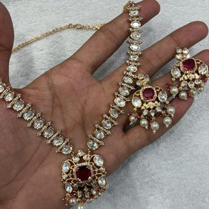 Puede incluir: Un conjunto de collar y pendientes en tono dorado con piedras transparentes de corte ovalado, piedras centrales de corte cuadrado en rojo oscuro y pequeñas cuentas blancas tipo perla. El collar tiene un colgante central y los pendientes hacen juego.