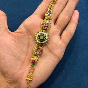 Pode incluir: Uma pulseira de tom dourado com um medalhão circular central contendo uma pedra verde-escura rodeada por pequenas contas brancas. A corrente é acentuada com quatro elos decorativos menores, cada um incrustado com pedras rosas e verdes.