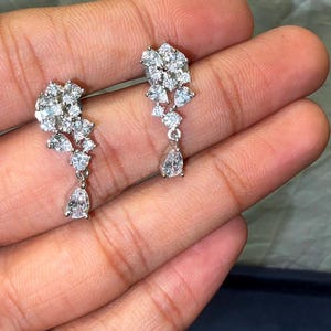May include: A pair of silver-toned drop earrings featuring a cluster of round, brilliant-cut clear stones arranged in a cascading floral-like pattern, terminating in a single pear-shaped drop stone suspended from the base of each earring.