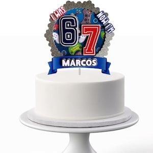 Puede incluir: Una tarta blanca glaseada con un adorno. El adorno presenta los números "6" y "7" con una combinación de colores azul y rojo, junto con el nombre "MARCOS" en una pancarta azul. El tema es de videojuegos.