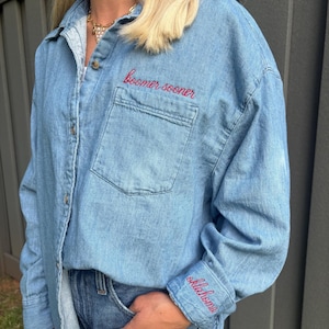 May include: A light blue denim button-down shirt featuring a chest pocket with the red embroidered text "boomer sooner" above it. The left sleeve cuff also displays the red embroidered text "oklahoma" in a cursive script font style.