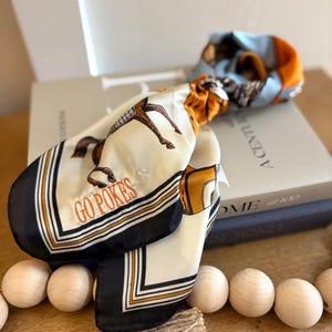 May include: A square silk scarf featuring a cream-coloured centre with a brown horse illustration and the orange text GO POKES. The scarf has a black border with white and orange stripes, and sections of light blue and orange fabric are visible.