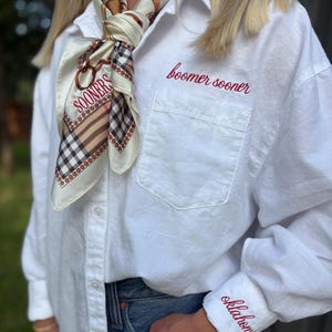May include: A white long-sleeve button-down shirt featuring red embroidered text that reads "boomer sooner" above the chest pocket and "oklahoma" on the cuff. A cream-colored silk scarf with a brown and black plaid pattern and the word "SOONERS" is tied around the neck.