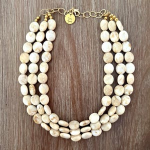 May include: A three-strand necklace featuring round, cream-colored beads with marbled brown patterns. The necklace includes a gold-toned metal chain, a lobster clasp, and a small circular gold tag engraved with the text "Crown Jewels".