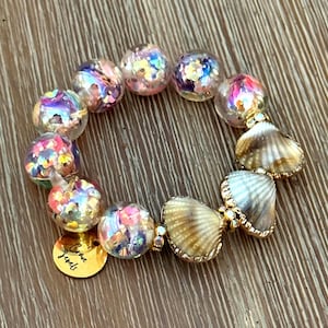 May include: A beaded bracelet featuring clear, iridescent beads filled with colorful confetti and seashell accents. A gold charm with the words "Crave Jewels" is attached. The bracelet is on a wooden surface.
