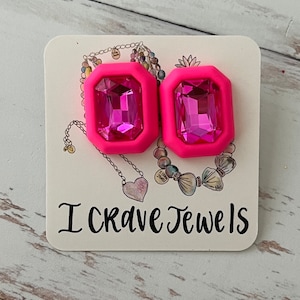 May include: A pair of pink statement earrings with large, faceted, pink gem-shaped stones. The earrings are on a white card with the text "I Crave Jewels".