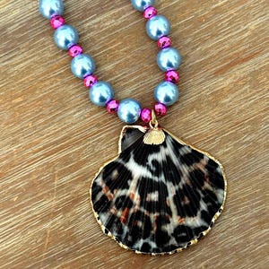 May include: A necklace featuring a large scallop shell pendant with a black and white leopard print pattern and a gold-toned rim. The strand consists of alternating light blue round beads and smaller, faceted hot pink beads in a repeating sequence.