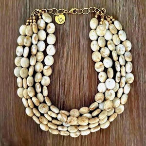 May include: A multi-strand necklace featuring oval-shaped, off-white beads with brown marbling. The necklace is connected by a gold-colored chain and clasp. A small, round gold charm with text is attached. The necklace is displayed on a wooden surface.