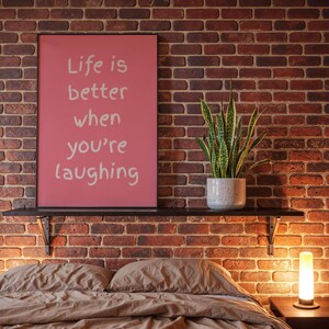 以下が含まれることがあります： ピンクの背景に白い文字で「Life is better when you're laughing」と書かれた長方形の額入りアート。黒い棚の上に飾られており、隣には斑点のあるライトグレーの陶器鉢に入った緑色のサンセベリアが置かれています。