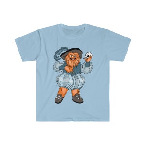 Op de afbeelding: Een lichtblauw t-shirt met een cartoonillustratie van een harige oranje schepsel dat is gekleed als een Shakespeare-personage en een schedel vasthoudt. Het schepsel heeft een brede, tandenrijke grijns en draagt een blauw en gouden kostuum.