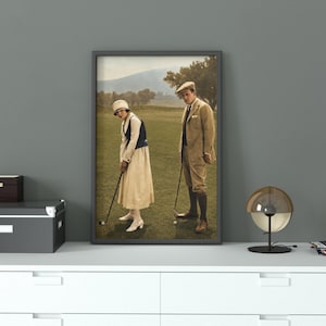 Puede incluir: Una fotografía vintage enmarcada muestra a dos personas jugando al golf en un campo verde. La persona de la izquierda lleva un vestido blanco y un sombrero, mientras que la de la derecha lleva un traje. Una caja negra y un baúl gris están sobre una superficie blanca.