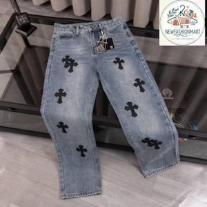 May include: Light blue denim jeans featuring a straight-leg cut, a button and zipper fly, and multiple black cross-shaped patches distributed across the front. A black brand tag is attached to the waistband with a small silver-toned metal charm.