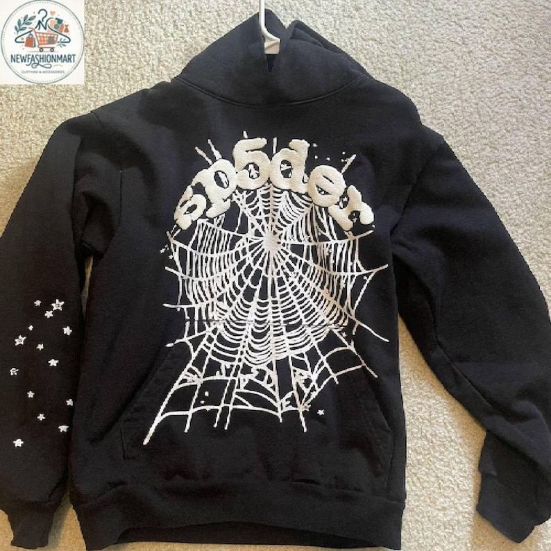 K&ouml;nnte beinhalten: Schwarzer Kapuzenpullover mit wei&szlig;em Spinnennetz-Design und dem Wort "Spider" in einer stilisierten Schriftart. Der linke &Auml;rmel hat ein Muster aus wei&szlig;en Sternen. Der Pullover hat eine Vordertasche.