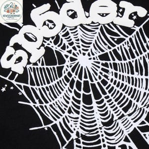 K&ouml;nnte beinhalten: Schwarzer Stoff mit einem wei&szlig;en Spinnennetz-Design und dem Wort "Spider" in einer aufgebl&auml;hten, wei&szlig;en Schriftart. Das Design enth&auml;lt kleine Sterne. Das Bild ist eine Nahaufnahme des Stoffes.