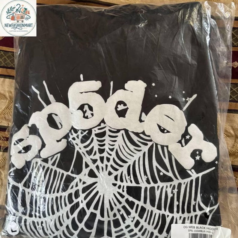 K&ouml;nnte beinhalten: Schwarzer Kapuzenpullover mit wei&szlig;em Spinnennetz-Design und dem Wort "spoder" in einer stilisierten Schriftart. Der Kapuzenpullover ist in einer transparenten Plastikt&uuml;te verpackt. Das Markenlogo befindet sich in der oberen linken Ecke.