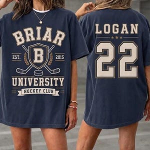 Może przedstawiać: Granatowy t-shirt z grafiką z przodu zawierającą napis BRIAR UNIVERSITY HOCKEY CLUB EST 2015 oraz tarczę z literą B, a także grafiką z tyłu z imieniem LOGAN i numerem 22 wykonanymi kremową czcionką w stylu uniwersyteckim.