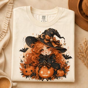 May include: A cream-colored t-shirt featuring a graphic of a character with long orange hair wearing a black witch hat with a bow and star accents. The design includes two jack-o'-lanterns, a black cat, bats, and autumn leaves.