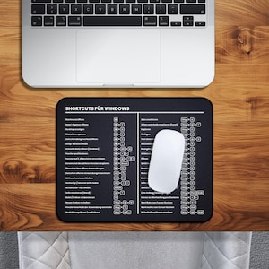 Könnte beinhalten: Ein schwarzes rechteckiges Mauspad mit weißem Text, der Windows-Shortcuts auflistet, und eine weiße kabellose Maus. Das Mauspad liegt auf einem Holztisch, im Hintergrund ist ein Laptop zu sehen. Der Text "SHORTCUTS FÜR WINDOWS" steht oben.